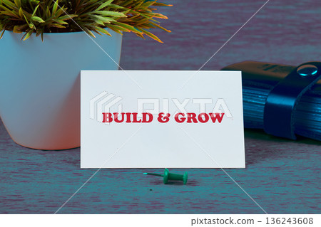 A white card displaying BUILD GROW in bold red text is positioned on a surface. A green pushpin rests below it A white card displaying BUILD GROW in bold red text is positioned on a surface. A green pushpin rests below it 136243608