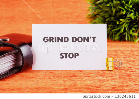 A white card reading GRIND DONT STOP stands on a textured orange surface, held by a yellow clip. A small dark book and green plant are nearby A white card reading GRIND DONT STOP stands on a textured orange surface, held by a yellow clip. A small dark book and green plant are nearby 136243611