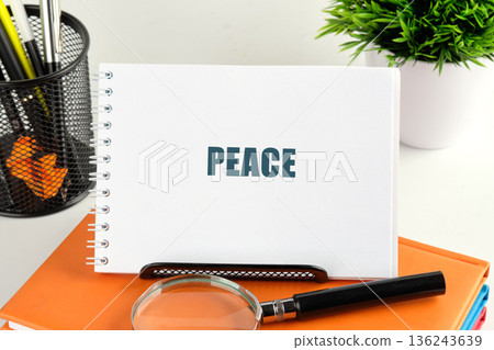 A white spiral notebook showing the word PEACE is on orange books. A magnifying glass, pen holder, and green plant complete the calm desk scene A white spiral notebook showing the word PEACE is on orange books. A magnifying glass, pen holder, and green plant complete the calm desk scene 136243639