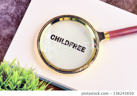 A gold-rimmed magnifying glass with a wooden handle rests on a white notebook, clearly enlarging the bold word CHILDFREE. Green foliage is visible on the side 136243650