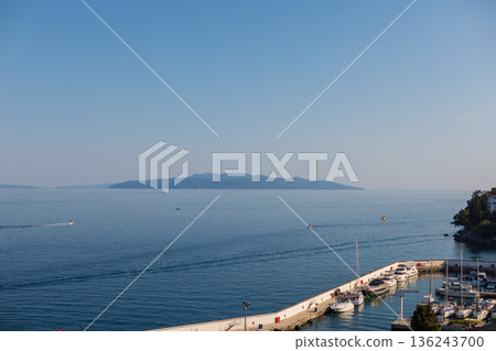 Tranquil Adriatic coast in Opatija, Croatia, with marina, pier, and moored boats against a backdrop of open sea and distant island on a sunny day. 136243700
