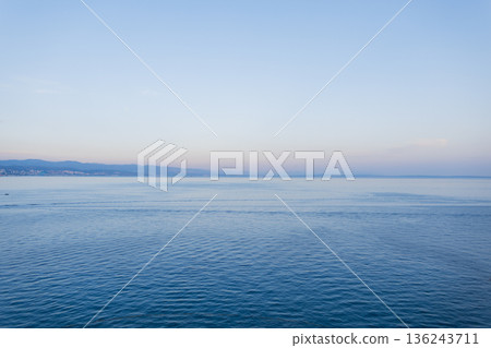 Adriatic Sea view near Opatija, Croatia, peaceful water with distant city and mountain range. Panoramic coastal landscape of Opatija, Croatia, with pastel sky, calm sea, and scenic mountains. 136243711