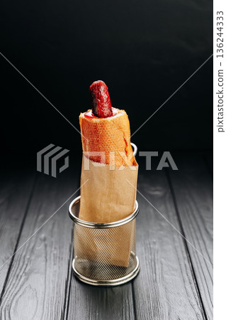 Creative hot dog in baguette with ketchup in paper bag on dark wooden background Creative hot dog in baguette with ketchup in paper bag on dark wooden background 136244333