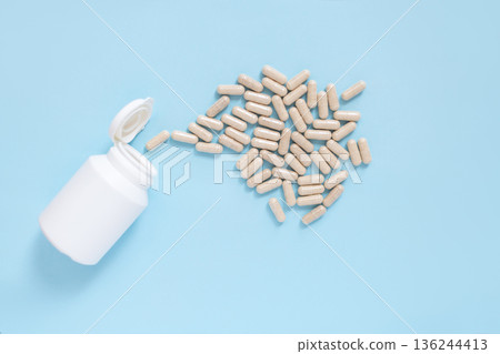 Dietary supplement capsules spilling from plastic container. Blue background with minimal style. Medicine, healthcare, or vitamins theme. Dietary supplement capsules spilling from plastic container. Blue background with minimal style. Medicine, healthcare, or vitamins theme. 136244413