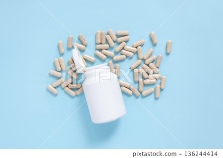 White plastic bottle with spilled beige capsules on a light blue background. Concept of medicine, healthcare, supplements, or vitamins. Clean, minimalistic composition for medical themes. 136244414