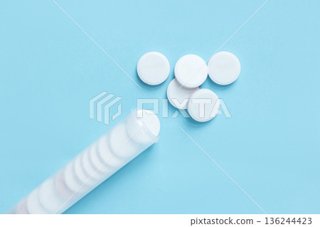 White round tablets scattered from a transparent plastic tube with a blue lid. Healthcare and pharmaceutical concept on a clean blue background White round tablets scattered from a transparent plastic tube with a blue lid. Healthcare and pharmaceutical concept on a clean blue background 136244423