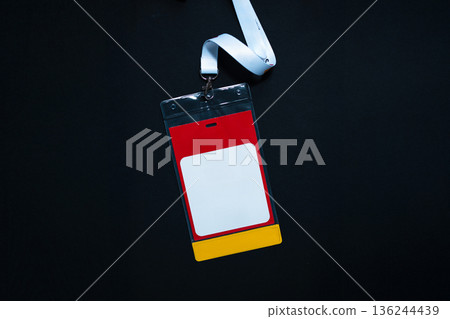 Blank name badge with red, white, and yellow design hanging on a white lanyard over black background. Concept of ID card, event pass, or conference badge. Free copy space for design 136244439