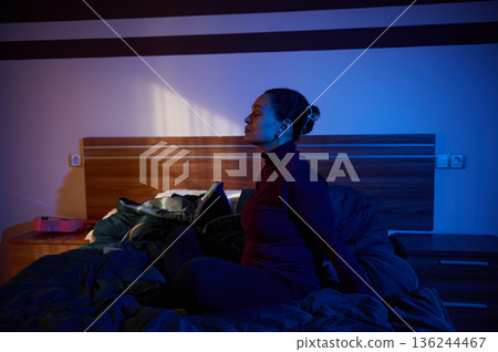 Relaxed Woman Sitting on Bed in Dim Blue Lighting For Quiet Night At Home 136244467
