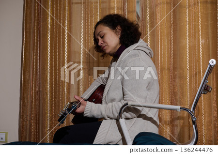 Woman Playing Acoustic Guitar While Sitting On A Bed In Cozy Apartment Setting Woman Playing Acoustic Guitar While Sitting On A Bed In Cozy Apartment Setting 136244476
