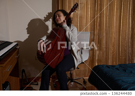 Woman Playing Acoustic Guitar In Cozy Home Studio With Relaxed Expression 136244480