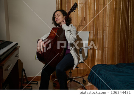 Relaxed Woman Plays Acoustic Guitar In Cozy Home Studio By Window And Bed Relaxed Woman Plays Acoustic Guitar In Cozy Home Studio By Window And Bed 136244481
