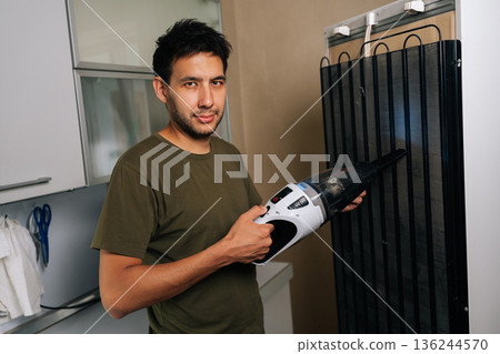Portrait of young man cleaning refrigerator back coils with handheld vacuum, removing dust buildup for improved cooling efficiency and lower energy consumption in modern kitchen setting 136244570