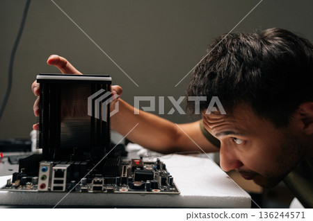 Close-up of computer technician male installing CPU cooler onto motherboard, performing maintenance and repair work on desktop computer in dark workshop. Concept of computer hardware, repairing. Close-up of computer technician male installing CPU cooler onto motherboard, performing maintenance and repair work on desktop computer in dark workshop. Concept of computer hardware, repairing. 136244571