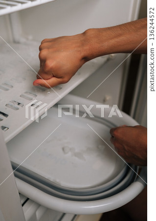 Vertical cropped shot of unrecognizable man squeezing melting ice cubes from refrigerator ice maker tray, hands wet from the task of defrosting freezer, water dripping frozen cubes removed. 136244572