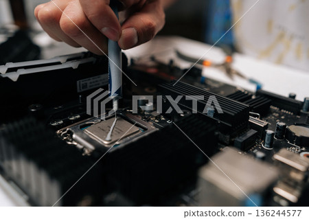 Technician hand applying thermal paste to CPU of computer motherboard, demonstrating process of computer maintenance and repair in dark workshop. Concept of computer hardware, repairing Technician hand applying thermal paste to CPU of computer motherboard, demonstrating process of computer maintenance and repair in dark workshop. Concept of computer hardware, repairing 136244577
