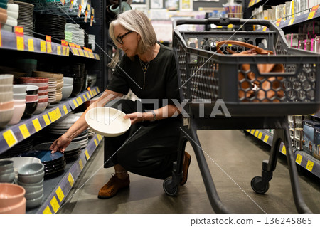 Woman shopping for bowls in a retail store with shopping cart and focused look on her face Woman shopping for bowls in a retail store with shopping cart and focused look on her face 136245865