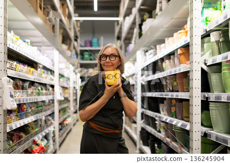Woman holds a yellow decorative pot in a store aisle filled with gardening supplies and pots 136245904