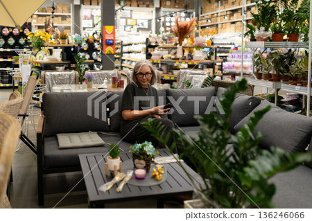 Woman sitting on a sofa in a store filled with plants and home decor enjoying her phone in a busy 136246066