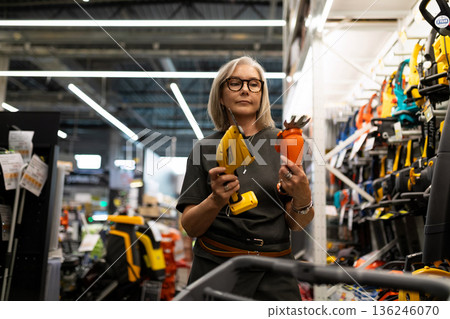 Woman shopping for tools in a hardware store while holding a glue gun and a decorative flower 136246070