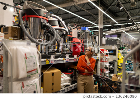 Woman shopping for cleaning equipment in a hardware store while considering options for a new vacuum 136246080