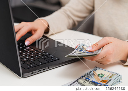 Female managing finances, counting cash and recording expenses on laptop. Savings, income tracking, and financial literacy. 136246688