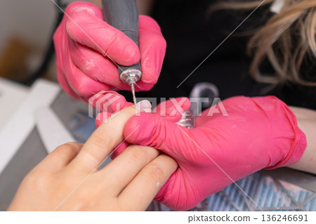Nail technician in pink gloves uses a drill to refine the nails during a manicure in a modern salon Nail technician in pink gloves uses a drill to refine the nails during a manicure in a modern salon 136246691