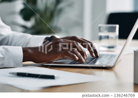 A professional Arab man sits at a desk in a modern office space, using a laptop. He types with concentration while documents and a glass of water are placed on the table in front of him. 136246709