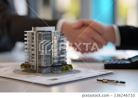 Two individuals shake hands above a model of a modern apartment building placed on a table. The scene suggests an agreement in the fields of architecture or real estate. Two individuals shake hands above a model of a modern apartment building placed on a table. The scene suggests an agreement in the fields of architecture or real estate. 136246710