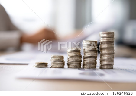 Coins are stacked in ascending order on a surface covered with papers. The workspace background shows a blurred figure engaged in an office task during daytime hours. Coins are stacked in ascending order on a surface covered with papers. The workspace background shows a blurred figure engaged in an office task during daytime hours. 136246826