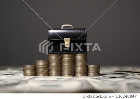 A miniature business briefcase is positioned on top of stacks of coins. Surrounding the briefcase are dollar bills laid out on a flat surface. This scene highlights themes of finance and business. 136246847