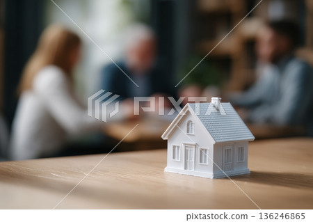 A small model house sits on a desk while two people discuss real estate in the background. This scene captures a business meeting focused on property investment. A small model house sits on a desk while two people discuss real estate in the background. This scene captures a business meeting focused on property investment. 136246865