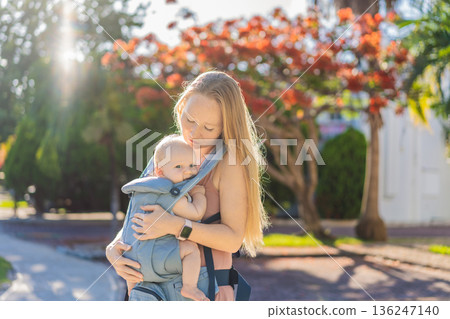 Mother using ergonomic baby carrier during a walk with her infant outdoors. Comfortable babywearing, active motherhood lifestyle, bonding, modern parenting, hands free care and everyday family routine 136247140