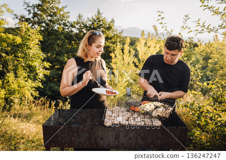Couple grilling vegetables outdoors at sunset, tasting food at summer barbecue Couple grilling vegetables outdoors at sunset, tasting food at summer barbecue 136247247