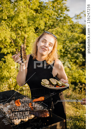 Smiling woman holding grilled vegetables and plate at outdoor barbecue in golden hour Smiling woman holding grilled vegetables and plate at outdoor barbecue in golden hour 136247256