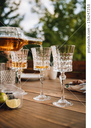 Pouring whiskey into crystal glass on wooden table, outdoor party with berries and bokeh Pouring whiskey into crystal glass on wooden table, outdoor party with berries and bokeh 136247258