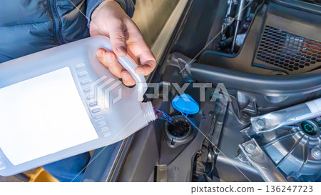 Person pouring oil from a plastic container into a car engine compartment, visible engine parts and tools in the background 136247723