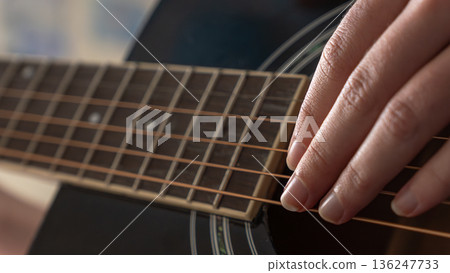 Close-up of a hand playing acoustic guitar strings, showcasing the fretboard and body of the instrument in a cozy indoor setting Close-up of a hand playing acoustic guitar strings, showcasing the fretboard and body of the instrument in a cozy indoor setting 136247733