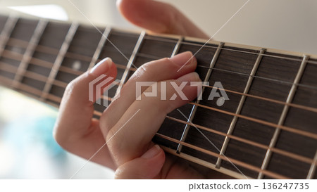 Close-up of hand pressing guitar strings on fretboard, showcasing finger positioning and wooden texture in a bright indoor setting Close-up of hand pressing guitar strings on fretboard, showcasing finger positioning and wooden texture in a bright indoor setting 136247735
