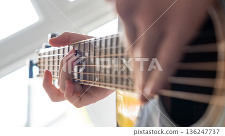 Close-up of a person playing an acoustic guitar, fingers pressing down on the strings, with sunlight streaming through a window Close-up of a person playing an acoustic guitar, fingers pressing down on the strings, with sunlight streaming through a window 136247737