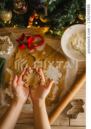 Holiday background. Female hands holding Christmas gingerbread snowflake cookie, cookie cutters and dough, top view, flat lay 136248886