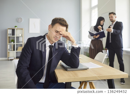 Frustrated businessman rubs forehead as coworkers discuss him behind him Frustrated businessman rubs forehead as coworkers discuss him behind him 136249432