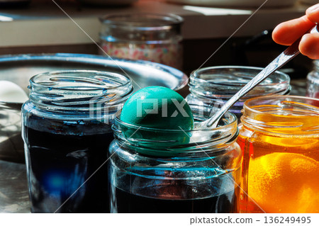 Hand holding spoon with dyed egg taking it out of glass jar during with blue food dye during easter egg coloring 136249495