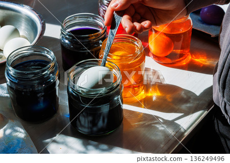 Sunlit close up of a hand using a spoon to dip a white egg into a blue food dye jar, with orange dye jars and other supplies on a kitchen table 136249496