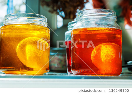 Close up of a easter egg in a yellow and orange food dye solution in a glass jars on a sunlit kitchen table Close up of a easter egg in a yellow and orange food dye solution in a glass jars on a sunlit kitchen table 136249500