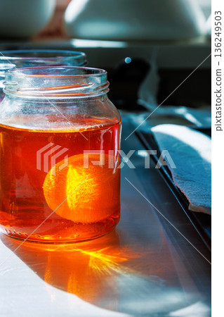 Egg soaking in orange food dye in glass jar on sunlit kitchen table 136249503