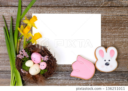 White card on a wooden background with daffodils, nest and Easter cookies. 136251011