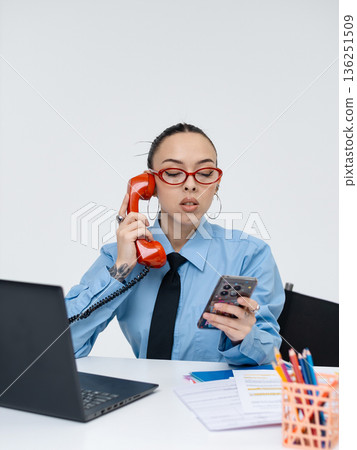 Young white woman in office attire working on laptop while talking on vintage red phone, isolated on white background Young white woman in office attire working on laptop while talking on vintage red phone, isolated on white background 136251509