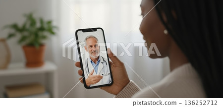 African american woman holding a smartphone and smiling, having a virtual online telemedicine consultation with a medical doctor discussing health on a video call from home 136252712