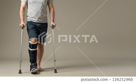 Man using crutches and wearing a medical orthopedic walking boot for a leg injury demonstrating empowerment and resilience during the rehabilitation process in a minimalist studio setting. 136252960
