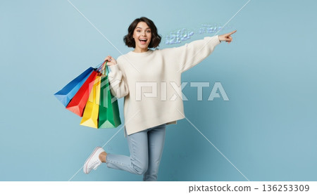 Cheerful young woman with colorful shopping bags pointing to copy space on blue background, celebrating retail therapy, sales, online checkout and happy consumer lifestyle 136253309